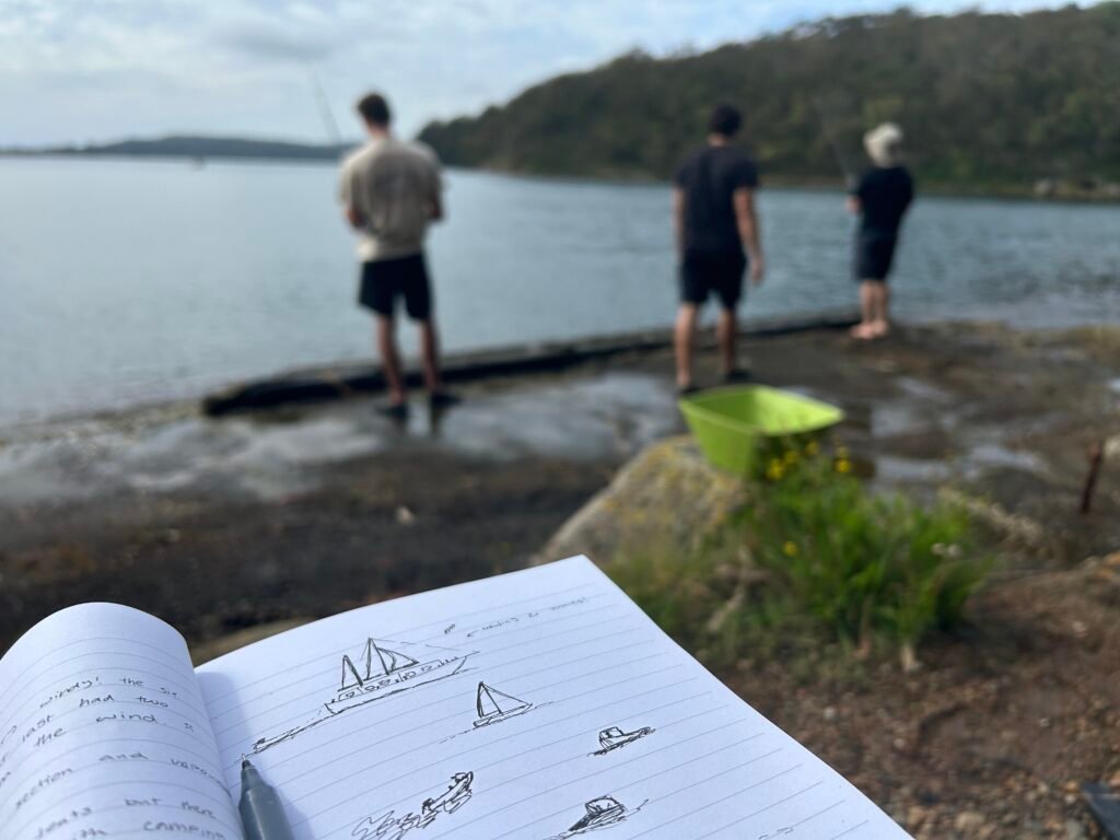 notebook full of drawings of boats with a blurry background containing three men fishing